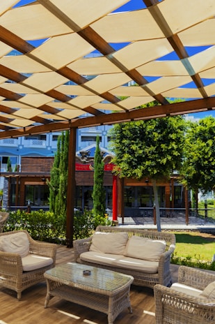 A cozy outdoor seating area featuring wicker furniture arranged under a wooden pergola with a crisscross pattern of beige canvas providing shade. The scene is set against a lush background of trees and shrubs, and a building with a modern facade is visible. Sunlight casts shadows across the furniture and the wooden floor.