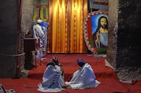 A religious setting featuring two people sitting on a red carpet, covered with white fabric, facing a large religious painting. In the background, a person dressed in white stands near a vibrant orange curtain with religious iconography on either side.