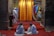 A religious setting featuring two people sitting on a red carpet, covered with white fabric, facing a large religious painting. In the background, a person dressed in white stands near a vibrant orange curtain with religious iconography on either side.