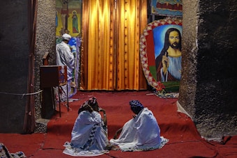 A religious setting featuring two people sitting on a red carpet, covered with white fabric, facing a large religious painting. In the background, a person dressed in white stands near a vibrant orange curtain with religious iconography on either side.