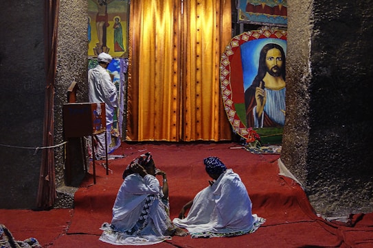 A religious setting featuring two people sitting on a red carpet, covered with white fabric, facing a large religious painting. In the background, a person dressed in white stands near a vibrant orange curtain with religious iconography on either side.