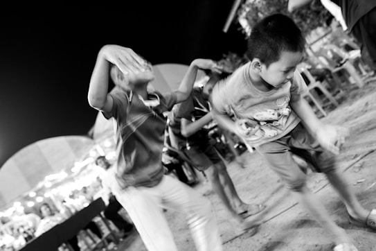 Children are energetically dancing outdoors at night, with blurred movement capturing the dynamism and joy of the scene. The background features a row of tables and chairs under festive lights, indicating a celebratory event.