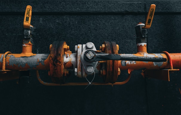 A close-up view of an industrial valve connected to orange and grey pipes. The valve mechanism is slightly rusted, with flaking paint and visible bolts. Two handles are attached on either side, with one prominently angled toward the viewer.