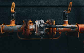 A close-up view of an industrial valve connected to orange and grey pipes. The valve mechanism is slightly rusted, with flaking paint and visible bolts. Two handles are attached on either side, with one prominently angled toward the viewer.