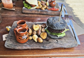 A rustic wooden platter features a black charcoal burger garnished with lettuce and topped with sesame seeds, alongside a serving of roasted potatoes. Two small decorative clay pots accompany the meal, adding a traditional touch.
