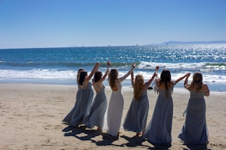 A group of women wearing long dresses stand in a line on a sandy beach, holding hands with their arms raised. The ocean waves gently roll in the background under a clear blue sky.
