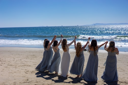 A group of women wearing long dresses stand in a line on a sandy beach, holding hands with their arms raised. The ocean waves gently roll in the background under a clear blue sky.