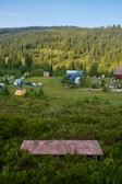 A serene campsite set amidst lush green hills and a dense forest. Several tents are scattered across a grassy field, with a few small buildings in the background. The sky is clear with a hint of blue, and the scene is peaceful and inviting.