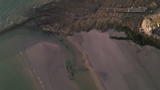 An aerial view of a coastline with sandy beaches, rocky areas, and parts of a building near the water. The sea is visible on the left, and there are textured patterns in the sand and rocks.
