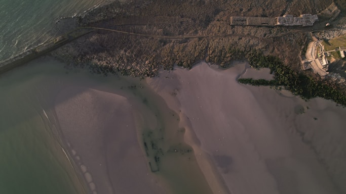 An aerial view of a coastline with sandy beaches, rocky areas, and parts of a building near the water. The sea is visible on the left, and there are textured patterns in the sand and rocks.