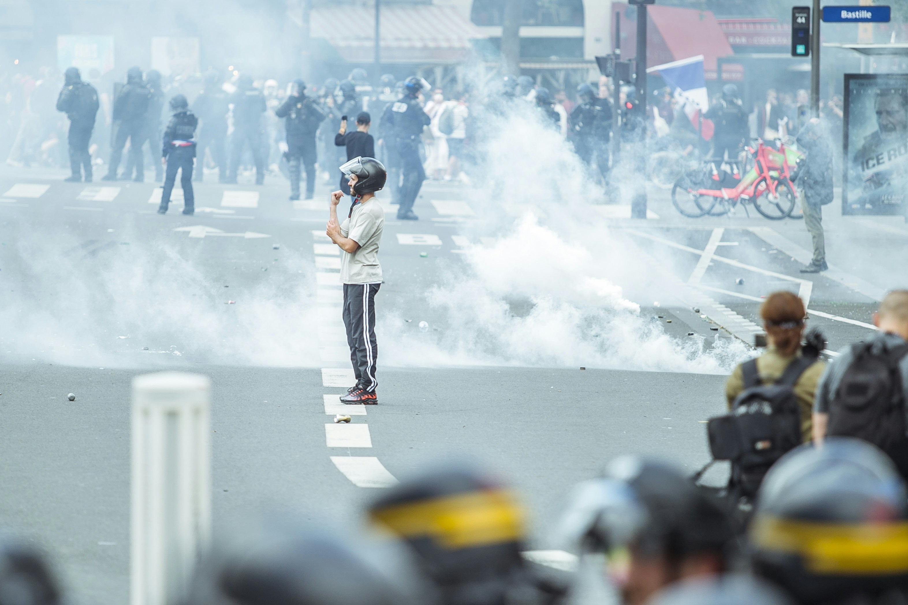 A young man wearing black pants, a white shirt and a black motorcycle helmet is standing in the middle of a city street surrounded by smoke. There are police in riot gear in both the fore and background. (civil unrest)