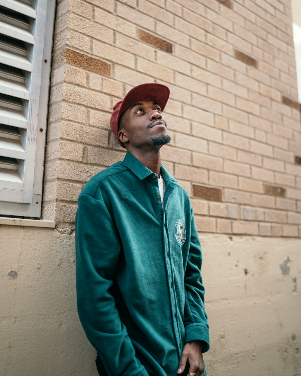 man in green zip up jacket and red cap standing beside brown brick wall during daytime