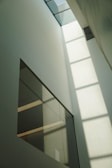 A tall, minimalist interior space with large windows that allow sunlight to cast geometric shadows on the walls. The design features clean lines and a muted color palette, creating a sense of openness and calm.