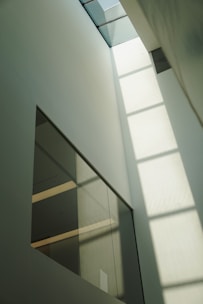 A tall, minimalist interior space with large windows that allow sunlight to cast geometric shadows on the walls. The design features clean lines and a muted color palette, creating a sense of openness and calm.