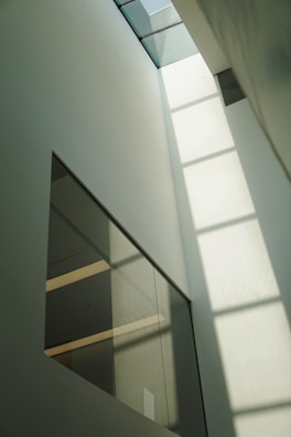 A tall, minimalist interior space with large windows that allow sunlight to cast geometric shadows on the walls. The design features clean lines and a muted color palette, creating a sense of openness and calm.