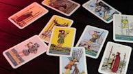 A collection of tarot cards scattered on a wooden surface, lit with a red light. Each card displays different figures and symbols, with illustrations showing various scenes and characters from the tarot deck.