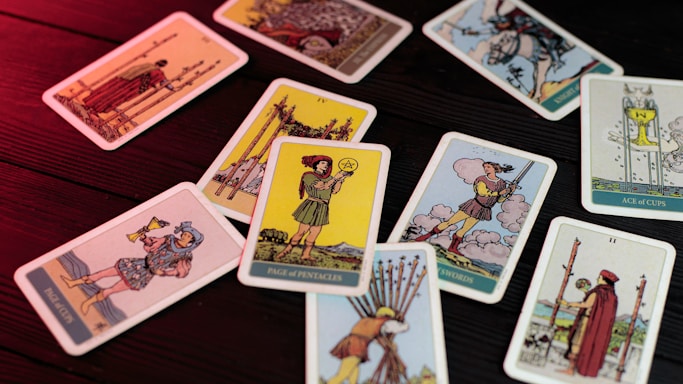 A collection of tarot cards scattered on a wooden surface, lit with a red light. Each card displays different figures and symbols, with illustrations showing various scenes and characters from the tarot deck.