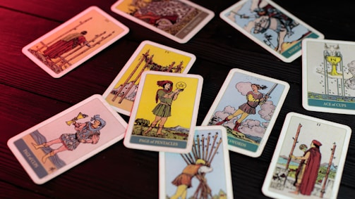 A collection of tarot cards scattered on a wooden surface, lit with a red light. Each card displays different figures and symbols, with illustrations showing various scenes and characters from the tarot deck.