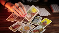 A hand with painted nails holds up a tarot card above a wooden table scattered with various other tarot cards. Some cards are face up, displaying intricate and colorful designs, while others are face down. The lighting is warm, creating a mystical and intimate atmosphere.