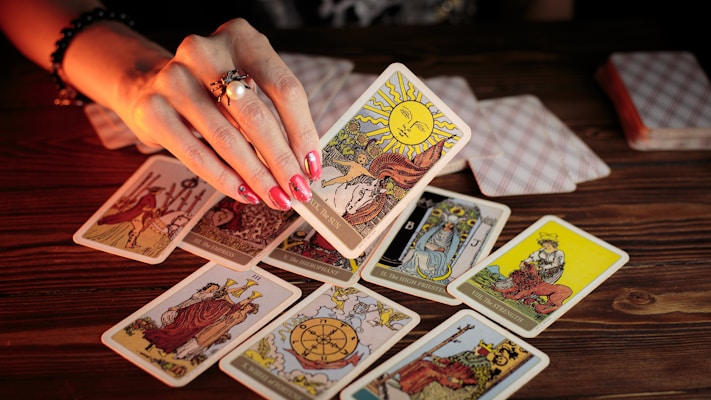 A hand with painted nails holds up a tarot card above a wooden table scattered with various other tarot cards. Some cards are face up, displaying intricate and colorful designs, while others are face down. The lighting is warm, creating a mystical and intimate atmosphere.