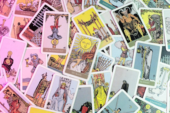 A collection of Tarot cards is scattered across a surface, displaying a variety of illustrations and symbolism. Some cards are upright, while others are upside down, featuring figures such as 'The Hanged Man,' 'The Sun,' 'The Fool,' and 'The Devil.' The cards are colorful and intricately designed, often depicting medieval or mythical scenes with characters, animals, and symbolic objects.