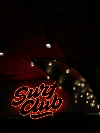 Neon sign in red with cursive text reading 'Surf Club' is illuminated against a dark background. Adjacent to the sign are exposed light bulbs hanging from a ceiling, casting a soft glow. The ceiling appears to be made of dark wood panels, enhancing the moody ambiance.