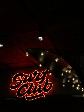 Neon sign in red with cursive text reading 'Surf Club' is illuminated against a dark background. Adjacent to the sign are exposed light bulbs hanging from a ceiling, casting a soft glow. The ceiling appears to be made of dark wood panels, enhancing the moody ambiance.