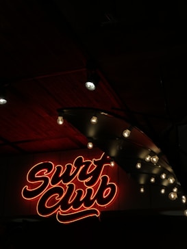 Neon sign in red with cursive text reading 'Surf Club' is illuminated against a dark background. Adjacent to the sign are exposed light bulbs hanging from a ceiling, casting a soft glow. The ceiling appears to be made of dark wood panels, enhancing the moody ambiance.