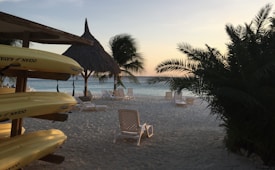 A serene beach scene with yellow kayaks stacked on the left side, palm fronds framing the view, and empty lounge chairs scattered across white sand. The ocean is visible in the background with a sky transitioning into sunset colors.