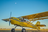 A yellow, small aircraft with visible landing gear is parked on a grassy field under a clear blue sky. The aircraft is labeled 'EXPERIMENTAL' on the side. There is a person in the cockpit wearing sunglasses. The moon is visible in the sky above the plane.