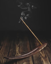 A burning incense stick placed in a wooden incense holder, with gentle wisps of smoke rising upwards against a dark background. The surface is a rustic, wooden texture, adding to the serene atmosphere.