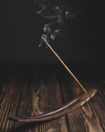 A burning incense stick placed in a wooden incense holder, with gentle wisps of smoke rising upwards against a dark background. The surface is a rustic, wooden texture, adding to the serene atmosphere.
