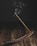 A burning incense stick placed in a wooden incense holder, with gentle wisps of smoke rising upwards against a dark background. The surface is a rustic, wooden texture, adding to the serene atmosphere.