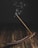 A burning incense stick placed in a wooden incense holder, with gentle wisps of smoke rising upwards against a dark background. The surface is a rustic, wooden texture, adding to the serene atmosphere.