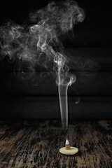 A cone-shaped incense burner sits on a wooden surface with smoke gracefully rising in swirling patterns against a dark background.
