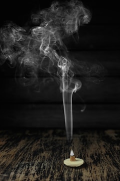 A cone-shaped incense burner sits on a wooden surface with smoke gracefully rising in swirling patterns against a dark background.