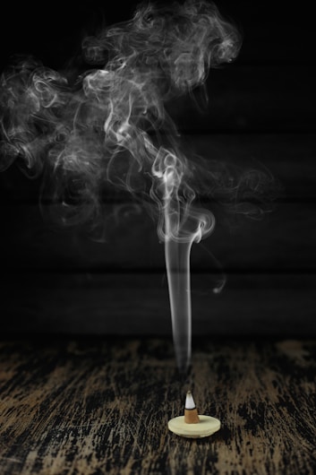 A cone-shaped incense burner sits on a wooden surface with smoke gracefully rising in swirling patterns against a dark background.
