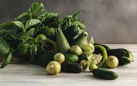 A collection of fresh green vegetables including basil leaves, zucchinis, cucumbers, Mexican husk tomatoes (tomatillos), and a green chili pepper arranged on a light wooden surface against a neutral background.