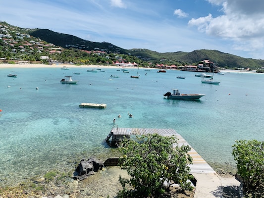 A clear turquoise bay dotted with several small boats floating peacefully. In the foreground, a small dock leads to the water, surrounded by lush green plants. The background features a sandy beach lined with colorful buildings and houses built into the hills. The sky is mostly clear with some fluffy clouds.