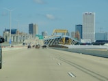 A multi-lane highway leads towards a city skyline featuring a distinctive yellow bridge structure. Several vehicles are traveling both towards and away from the urban area. High-rise buildings, including one with distinctive latticework, stand prominently against a clear, blue sky.