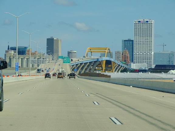 A multi-lane highway leads towards a city skyline featuring a distinctive yellow bridge structure. Several vehicles are traveling both towards and away from the urban area. High-rise buildings, including one with distinctive latticework, stand prominently against a clear, blue sky.