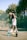 A shirtless person wearing a headband and shorts stands on a tennis court next to a tennis racket and ball. Two bottles are placed nearby, one filled with blue liquid and the other a black bottle. The court is surrounded by a chain-link fence and dense, green foliage.