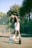 A shirtless person wearing a headband and shorts stands on a tennis court next to a tennis racket and ball. Two bottles are placed nearby, one filled with blue liquid and the other a black bottle. The court is surrounded by a chain-link fence and dense, green foliage.