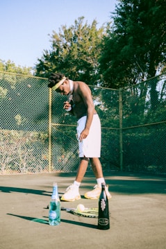 A shirtless person wearing a headband and shorts stands on a tennis court next to a tennis racket and ball. Two bottles are placed nearby, one filled with blue liquid and the other a black bottle. The court is surrounded by a chain-link fence and dense, green foliage.