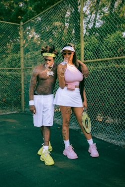 Two individuals stand on a tennis court with a chain-link fence in the background. The man is shirtless, wearing white shorts and bright yellow sneakers, with a headband, jewelry, and tattoos visible. The woman is dressed in a pink top, white skirt, and pink sneakers, holding a tennis racket and a glass. Her outfit is accessorized with sunglasses and a visor, and she has tattoos on her arms and legs.