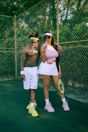 Two individuals stand on a tennis court with a chain-link fence in the background. The man is shirtless, wearing white shorts and bright yellow sneakers, with a headband, jewelry, and tattoos visible. The woman is dressed in a pink top, white skirt, and pink sneakers, holding a tennis racket and a glass. Her outfit is accessorized with sunglasses and a visor, and she has tattoos on her arms and legs.