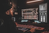 A person wearing a beanie is seated at a desk, working on digital audio workstations displayed on multiple computer monitors. The room appears to be a studio with soundproof walls, and the environment is dimly lit with a warm ambiance from overhead lighting. The screens show complex audio editing software with various tracks and controls.