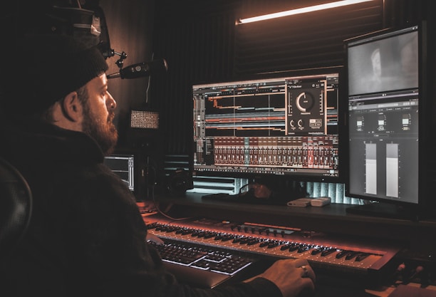 A person wearing a beanie is seated at a desk, working on digital audio workstations displayed on multiple computer monitors. The room appears to be a studio with soundproof walls, and the environment is dimly lit with a warm ambiance from overhead lighting. The screens show complex audio editing software with various tracks and controls.