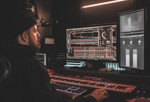A person wearing a beanie is seated at a desk, working on digital audio workstations displayed on multiple computer monitors. The room appears to be a studio with soundproof walls, and the environment is dimly lit with a warm ambiance from overhead lighting. The screens show complex audio editing software with various tracks and controls.