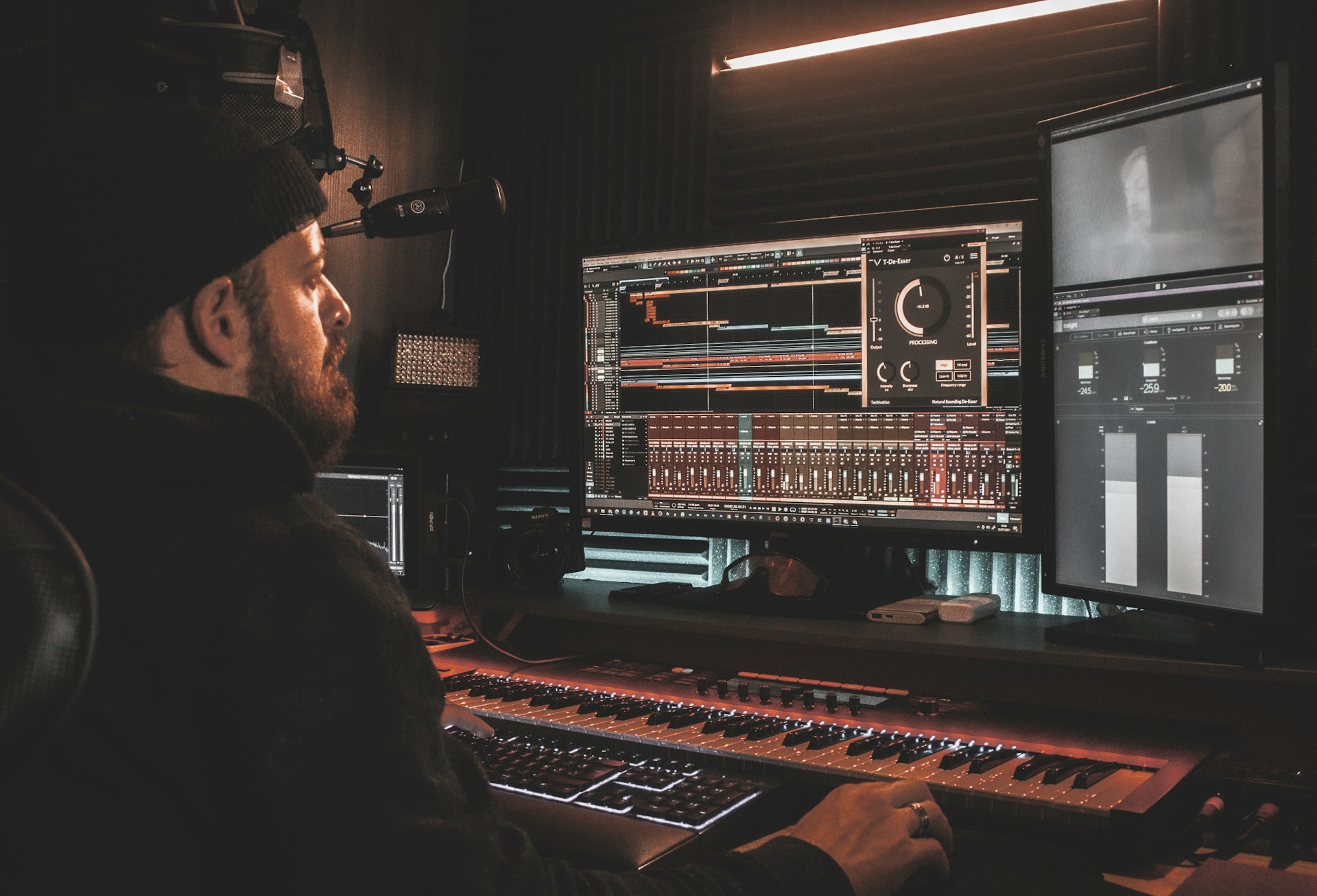 A person wearing a beanie is seated at a desk, working on digital audio workstations displayed on multiple computer monitors. The room appears to be a studio with soundproof walls, and the environment is dimly lit with a warm ambiance from overhead lighting. The screens show complex audio editing software with various tracks and controls.